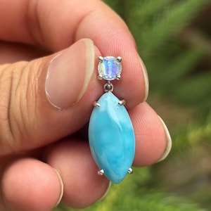 May include: A silver earring with a light blue, oval gemstone and a smaller, round, iridescent stone. The earring is held against a blurred green background. The oval stone has a pearlescent sheen.
