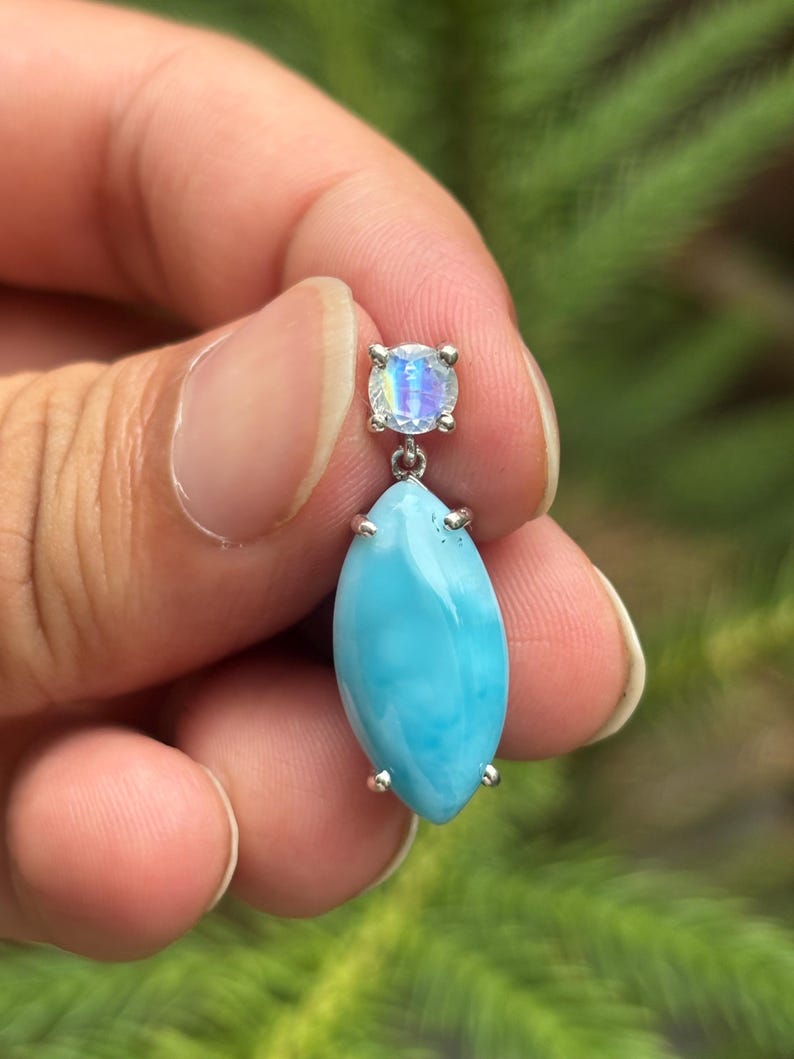 May include: A silver earring with a light blue, oval gemstone and a smaller, round, iridescent stone. The earring is held against a blurred green background. The oval stone has a pearlescent sheen.