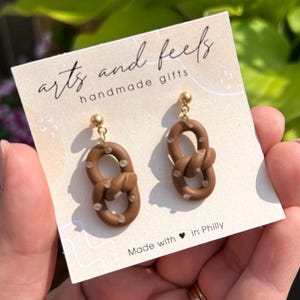 Op de afbeelding: Bruine pretzelvormige oorbellen met witte stippen, bevestigd aan goudkleurige pinnen. De oorbellen worden getoond op een kaart met de tekst "arts and feels handmade gifts" en "Made with in Philly."