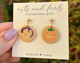 Könnte beinhalten: Ein Paar runde, hellbraune Ohrringe auf einer Präsentationskarte mit der Aufschrift 'arts and feels handmade gifts' und 'Made with heart in Philly'. Ein Ohrring zeigt ein violettes Geistermotiv, der andere einen orangefarbenen Kürbis.
