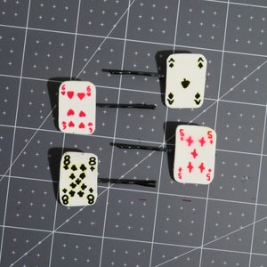 May include: Four white playing card hair clips with black and red details. The cards are the 6 of hearts, 8 of clubs, 5 of diamonds, and the ace of spades. The hair clips are attached to black bobby pins.