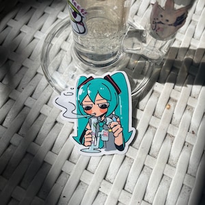 May include: A clear glass bong with a sticker of a teal-haired anime character smoking. The sticker is on a white surface. Other stickers are visible on the bong. The bong has a handle and a circular base.