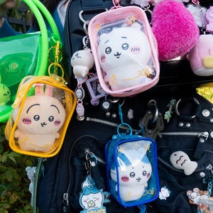 May include: A collection of colorful, clear bags and keychains featuring plush bunny characters. The bags are in green, yellow, and blue, with the keychains attached to a black backpack. Other accessories include a pink pom-pom and small plush figures.