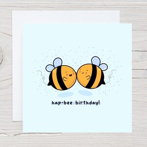 May include: A square greeting card with a light blue background features two cartoon bees with black and yellow stripes, blue wings, and smiling faces. The text "hap-bee birthday!" is written below the bees.