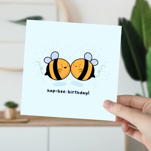 May include: A light blue square greeting card featuring two cartoon bees with black and yellow stripes, blue wings, and smiling faces. The text "hap-bee birthday!" is written below the bees.
