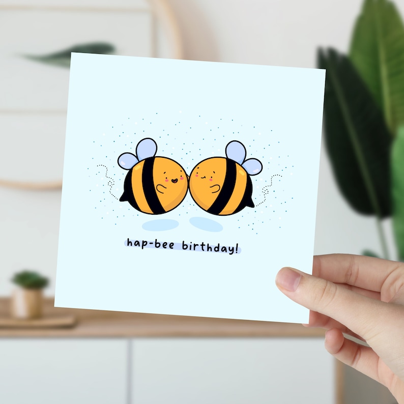 May include: A light blue square greeting card featuring two cartoon bees with black and yellow stripes, blue wings, and smiling faces. The text "hap-bee birthday!" is written below the bees.