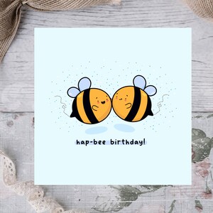 May include: A square greeting card with a light blue background features two cartoon bees with yellow and black stripes, blue wings, and smiling faces. The text "hap-bee birthday!" is written below the bees. The card is decorated with small blue dots.