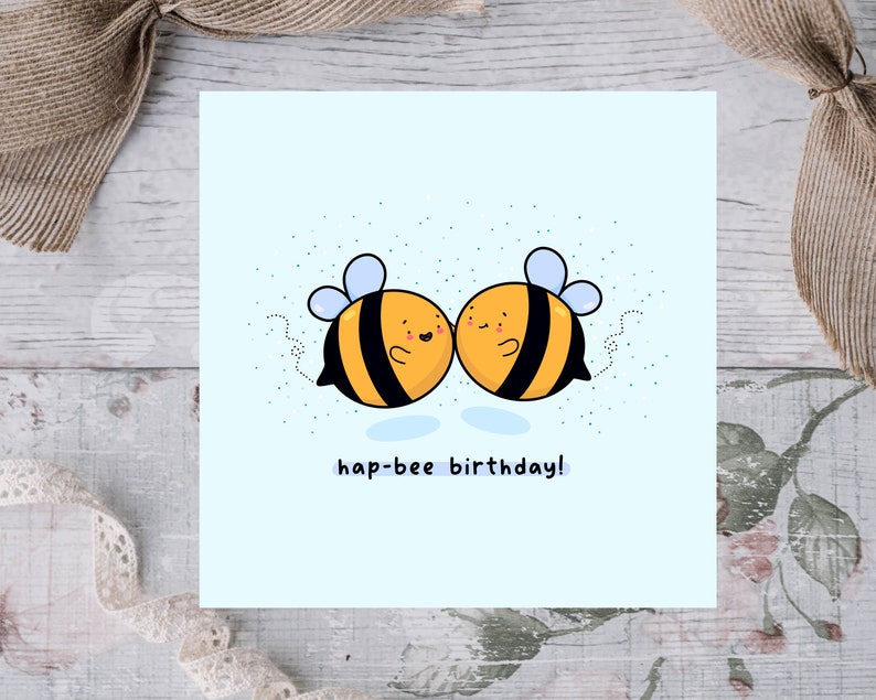 May include: A square greeting card with a light blue background features two cartoon bees with yellow and black stripes, blue wings, and smiling faces. The text "hap-bee birthday!" is written below the bees. The card is decorated with small blue dots.