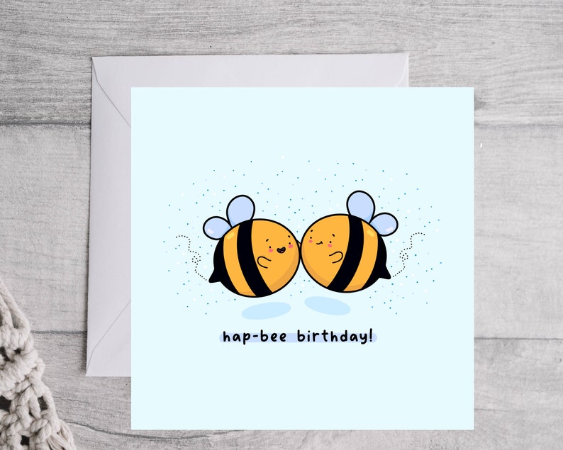 May include: A square greeting card with a light blue background features two cartoon bees with yellow and black stripes, blue wings, and smiling faces. The text "hap-bee birthday!" is written below the bees. The card is inside a white envelope.