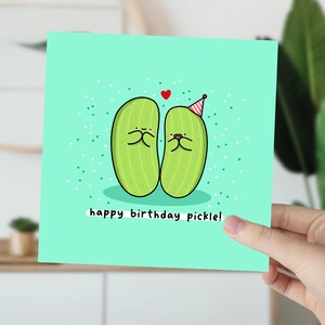 May include: A light green birthday card with two cartoon pickles wearing party hats. The card has a light blue background with white polka dots and the text "happy birthday pickle!"