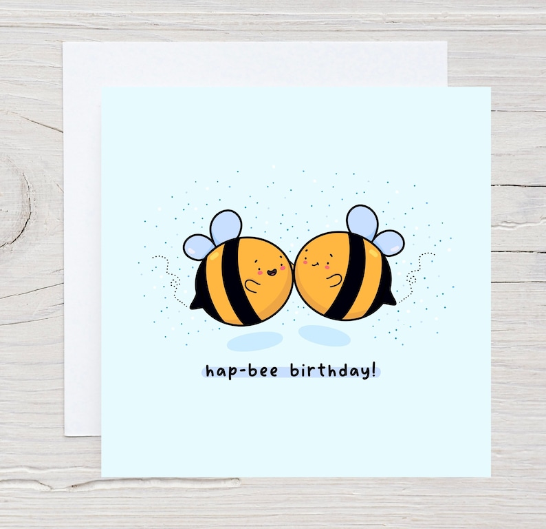 May include: A square greeting card with a light blue background features two cartoon bees with black and yellow stripes, blue wings, and smiling faces. The text "hap-bee birthday!" is written below the bees.
