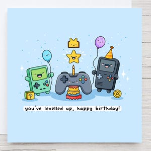 Könnte beinhalten: Hellblaue Geburtstagskarte mit einer Cartoon-Illustration von Videospielkonsolen, einem Controller und einem Kuchen. Der Text "you've levelled up, happy birthday!" steht unten auf der Karte.
