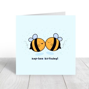 May include: A light blue greeting card featuring two cartoon bees with black and yellow stripes, smiling and touching. The text "hap-bee birthday!" is written below the bees. Small blue dots are scattered around the bees.