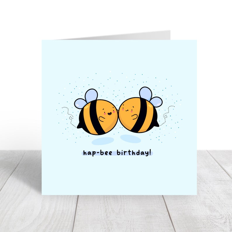 May include: A light blue greeting card featuring two cartoon bees with black and yellow stripes, smiling and touching. The text "hap-bee birthday!" is written below the bees. Small blue dots are scattered around the bees.