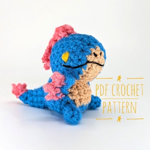 Può includere: Una creatura all'uncinetto fatta a mano in blu, rosa e beige. Il peluche ha un corpo blu, dettagli rosa e una pancia beige. Il testo "PDF CROCHET PATTERN" è scritto in giallo.