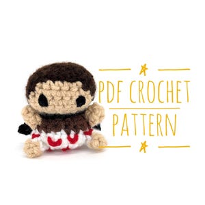 May include: A crocheted doll with a brown head, tan face, and black eyes. The doll has a brown and tan torso with red and white accents. The image includes the text "PDF CROCHET PATTERN" in yellow.