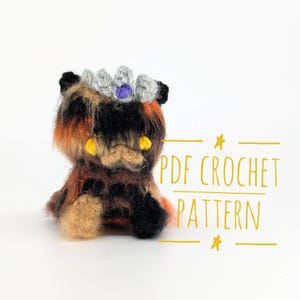 May include: A crocheted cat toy in shades of brown, orange, and black, wearing a silver crown with a purple jewel. The cat has yellow eyes. The image includes the text "PDF CROCHET PATTERN".