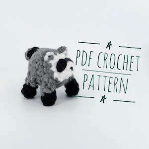 May include: A small, handmade crochet raccoon figurine featuring grey, white, and black yarn. The raccoon has black paws and a distinct facial mask. To the right of the figurine, the text "PDF CROCHET PATTERN" is written in dark green ink.