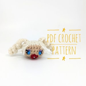 May include: A handmade crochet doll head with a light beige face, white hair, and blue eyes. The doll has a red mouth and is lying on a white surface. The text "PDF CROCHET PATTERN" is written in yellow.