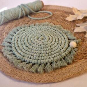 May include: A green crocheted coaster with a fringe border sits on a natural fibre placemat. The coaster has a circular design with a centre circle and concentric rings.