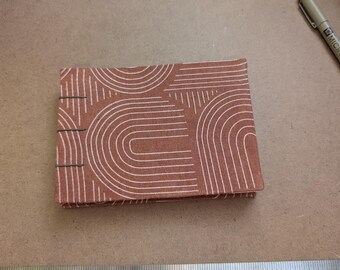 May include: A small, rectangular notebook with a terracotta-colored cover featuring a white, arched pattern. The notebook is closed and lies on a light brown surface. A metal ruler and pen are also visible.
