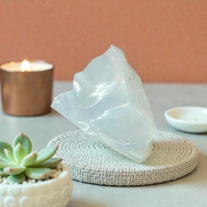 May include: A large, translucent crystal sits on a round, woven coaster. A lit candle in a copper-coloured holder and a small succulent in a white pot are in the foreground. A small white dish is in the background.