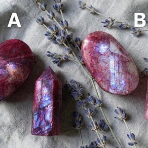 May include: Two polished, oval-shaped stones and two crystal points in shades of pink and purple, with iridescent flashes. The crystals are arranged with sprigs of lavender on a grey fabric background. The letter "A" is above the two crystals on the left, and the letter "B" is above the two crystals on the right.