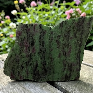 Pode incluir: Uma pedra em bruto com uma cor de base verde, acentuada por inclusões minerais pretas e vermelhas escuras. A pedra tem uma forma e textura natural e irregular, e é exibida em uma superfície de madeira com um fundo floral desfocado.