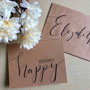 May include: Two brown cards with black calligraphy. One card says "Happy Birthday!" and the other says "Elizabeth". White flowers are in the upper left corner. The cards are on a light-coloured surface.