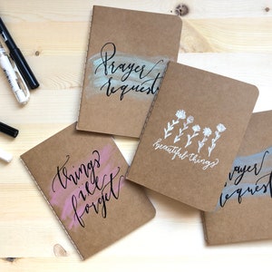 May include: Four notebooks with brown kraft paper covers, each featuring handwritten calligraphy. One says "Prayer Request" in black ink over a light blue brushstroke. Another says "beautiful things" with floral illustrations. The third says "things I'll forget" over a pink brushstroke.