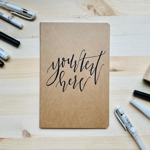 May include: A rectangular notebook with a light brown kraft paper cover featuring the black cursive text "your text here" centred on the front. The notebook is surrounded by various black and white pens and a small square wooden stamp.