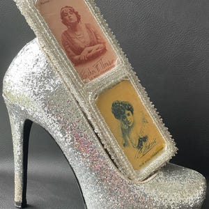May include: A silver glitter high heel shoe with a platform sole, holding two vintage cigarette cards. The cards feature portraits of women and the text "Fanchon T. Thomkins" and "Old Mill Cigarettes."
