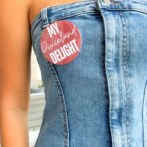 May include: A denim strapless top with a round, red button pinned to the front. The button has white text that reads "MY Dixieland DELIGHT". The top is light blue and has visible seams.