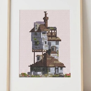 May include: A cross-stitch pattern of a multi-story house with a tall chimney, brown roof sections, and gray walls, displayed in a light wood frame. The design features intricate pixelated details, small windows, and green foliage at the base.