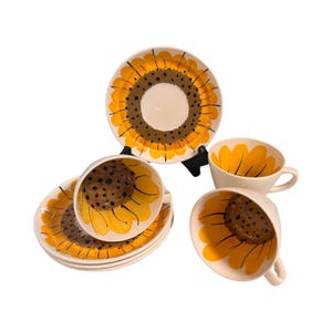 Könnte beinhalten: Ein Satz Keramik-Teetassen und Untertassen mit Sonnenblumenmuster. Die Tassen und Untertassen sind weiß mit gelben und braunen Blumenmustern. Die Tassen haben Henkel und die Untertassen sind rund.