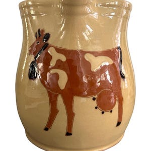 May include: A beige ceramic pitcher featuring a hand-painted brown and cream cow illustration on the front. The cow is depicted in profile, wearing a black collar with a bell, and the pitcher has a wide, curved rim with a central pouring spout.