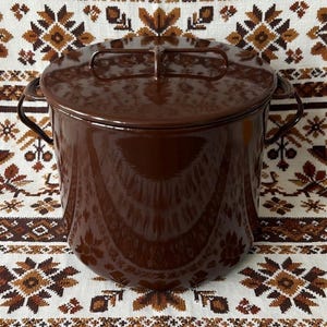 Puede incluir: Una olla grande de esmalte marrón con tapa a juego y asas laterales. La olla se coloca sobre un fondo de tela estampada con diseños florales marrones y blancos. Es un artículo de menaje.