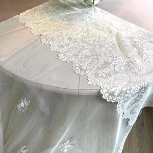 May include: A sheer white lace table runner with intricate floral and paisley patterns. The runner has a scalloped edge and is draped over a round table. Delicate floral accents are scattered across the sheer fabric.