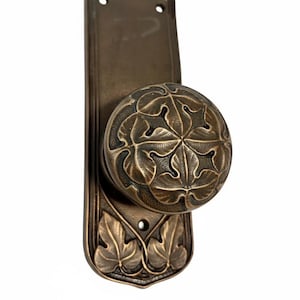 May include: Bronze-toned door knob and backplate with a detailed leaf design. The round knob features an intricate pattern of leaves, while the backplate has a vertical shape with leaf accents. The hardware is likely for a door.