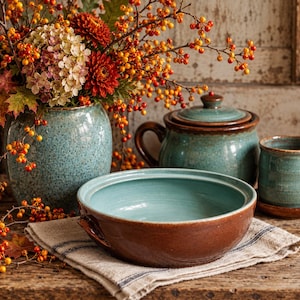May include: A ceramic collection featuring a speckled teal vase filled with autumn flowers, a lidded teal and brown pot, a matching teal mug on a saucer, and a wide, shallow teal and brown bowl resting on a striped linen cloth.