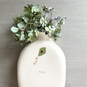 May include: White ceramic vase shaped like a bottle with a green kite with a red heart painted on it. The vase has the word "free" written on it. Dried green flowers are in the vase.