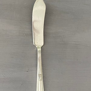 May include: A silver butter knife with a curved blade and a long, ridged handle. The handle has a decorative design near the base and an engraved monogram. The knife is set against a gray background.