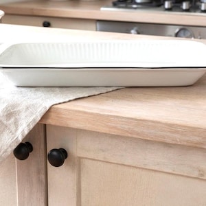 May include: A white rectangular baking dish with a black trim sits on a wooden countertop. The dish has a ribbed interior and is next to a beige linen towel. A bowl of fruit and a loaf of bread are in the background.