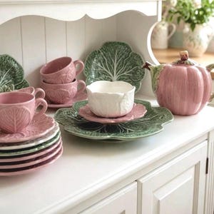 May include: A collection of ceramic tableware featuring pink and white cups, saucers, and a pumpkin-shaped teapot, alongside green cabbage-leaf patterned plates. The set includes stacked pink saucers and a white bowl on a green plate.