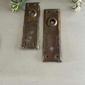 May include: Two antique brass door plates with keyholes and rounded edges. The plates are slightly tarnished and have a rustic look.