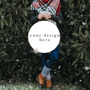 May include: A person holding a white round sign with the text "your design here" in black lettering. The sign is in front of a green evergreen tree with snow falling.