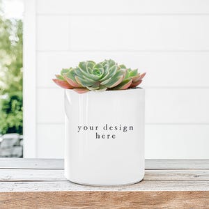 Puede incluir: Una maceta de cerámica blanca con el texto "your design here" contiene una suculenta verde con bordes rojizos. La maceta está sobre una superficie de madera, con una pared blanca y vegetación en el fondo.