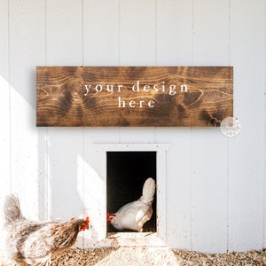 May include: A white wooden coop with a brown wooden sign that says "your design here" above the entrance. Two chickens are near the coop, one white chicken is inside the coop and one gray chicken is outside the coop.