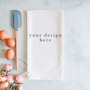 May include: A white kitchen towel with the text "your design here" printed on it. The towel is lying flat on a marble surface with three brown eggs, a blue spatula with a wooden handle, and pink flowers.