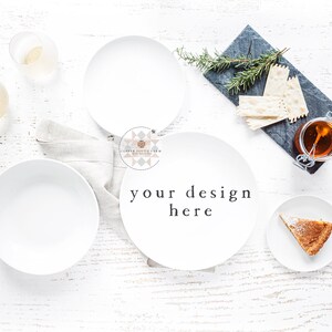 Op de afbeelding: Een witte tafelsetting met twee witte borden, een witte kom, twee glazen, een klein bord met een stuk taart, een leisteen serveerschaal met crackers en een pot honing, en een takje rozemarijn.